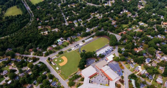 Walter M. Williams High School in Burlington, NC is seen below us as we fly over. The school building complex is in front and we also see a baseball field, a football field with bleachers.