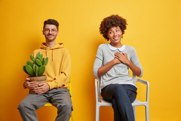 Fototapeta premium Happy pleased woman makes thankful gesture glad to receive cactus in pot from husband. Family couple in casual clothes sit on chairs isolated on yellow background. Domestic lifestyle concept