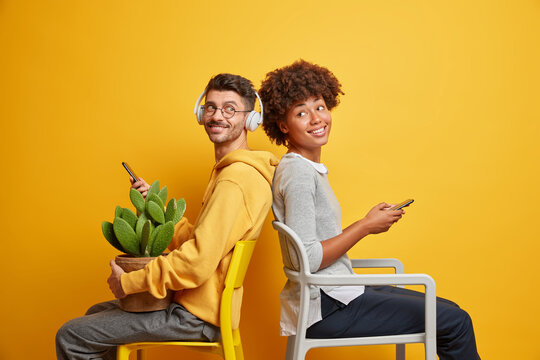 Positive teenage female and male addicted to modern technologies sit back to each other on chairs hold mobile phones dressed casually enjoy online communication isolated over yellow studio wall