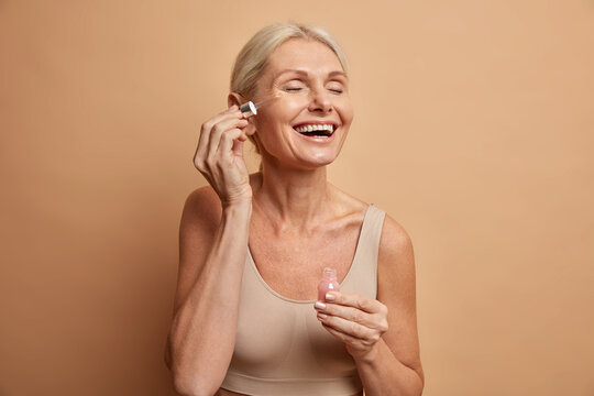 Happy senior lady applies cosmetic oil serum on face takes care of skin and smiles broadly enjoys beauty treatments stays always young beautifull poses in casual top against beige background