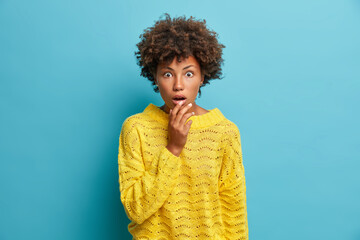 Cute amazed curly haired woman finds out shocking news stares at camera with wide opened mouth stands shocked and astonished wears casual yellow jumper isolated on blue background. Human reaction