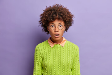 Portrait of amazed speechless woman with curly hair stares shocked impressed keeps mouth wide opened wears round glasses and casual green jumper isolated over purple background. Omg concept.