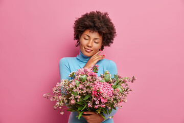 Pretty affectionate woman touches gently jawline stands with closed eyes holds big bunch of flowers dressed in casual blue jumper gets bouquet as gift from husband isolated on pink background