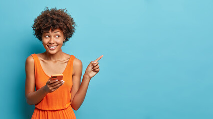Nice attractive young Afro American woman uses cell phone and points on copy space shows direction...