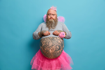 Photo of obese displeased gloomy bearded man fairy with wings holds magic wand and looks unhappily...