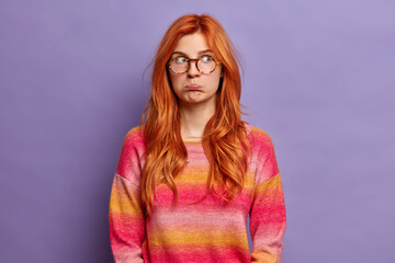 Frustrated displeased offended redhead European woman purses lips and feels insulted has bad mood depression dressed casually isolated on purple studio background. Negative emotions concept.