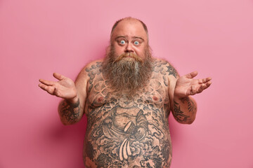 Hesitant fatso man spreads palms in clueless gesture has thick beard has shirtless fat tattoed body...