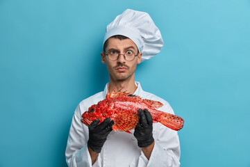 Photo of serious male cook carries fish looks directly at camera asks chef for advice what better to prepare tries tasty yummy recipe wears white uniform isolated on blue background. Cooking concept