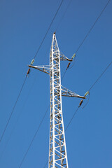 High voltage pole and electricity transmission line on the background of a slope with trees and sky.