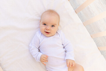 portrait of a small baby girl 6 months old in a white bodysuit lying on her back on a white bed, space for text