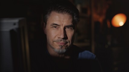 Obraz premium Facial portrait of mature man in his 50s at home sitting in dark living room.