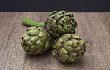 Three fresh organic purple artichokes with stem accents on a rustic wooden surface and dark...