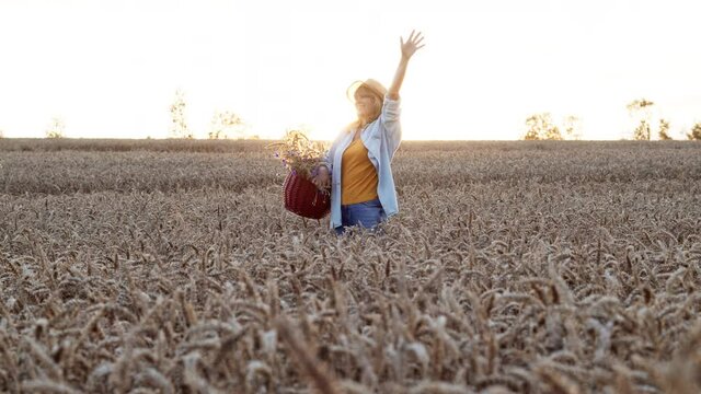 Woman With Basket Of Flowers Walks Field With Wheat. Romantic Image Of Blonde In Hat At Sunrise. She Shouts And Waves As If Greeting Or Calling Someone.