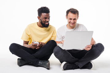Two mixed race men with phone and laptop sitting on the floor isolated on white background