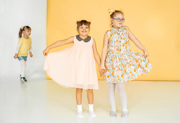 Two little girls in dresses are dancing in the studio on a yellow background. A child with bows on...