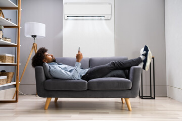 Young Man Lying Under Air Conditioner On Couch Using Smartphone