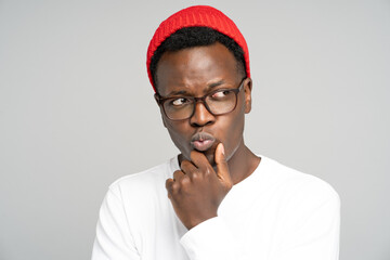 Studio portrait of unsure doubtful Black man in red hat wear glasses holds chin, looking right doubtfully, plans or reconsiders something, feeling hesitant poses against white background, copy space. 