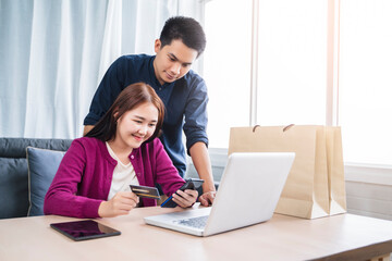 Asian couple online shopping ecommerce together from home modern living room using phone computer laptop tablet payment paying transaction money transfer credit debit card smiling happy joyfully