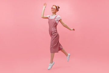 Red-haired woman in overalls and glasses joyfully moves on pink background