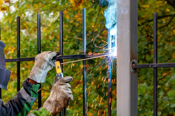 A welder in a protective helmet and gloves works with metal, welds a metal fence against a blurred background of autumn greenery.