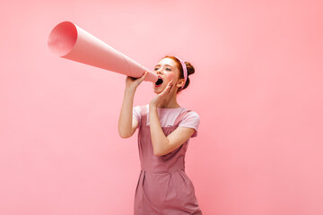 Cheerful woman with buns screams into mouthpiece. Girl in overalls posing on pink background