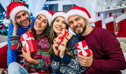 Group of beautiful best friends in Santa hats with gifts in hands are posing and having fun on Christmas tree background at home party