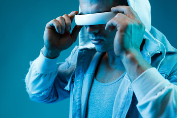 Young man in denim jacket on dark background. Guy using VR helmet. Augmented reality, virtual reality, future technology, game concept. Blue neon light.