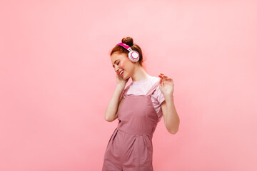 Positive redhead lady in pink outfit laughs while listening to music with pink headphones on isolated background