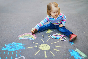 Fototapeta premium Adorable toddler girl drawing with colorful chalks on asphalt