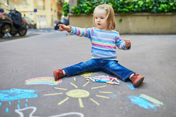 Naklejka premium Adorable toddler girl drawing with colorful chalks on asphalt