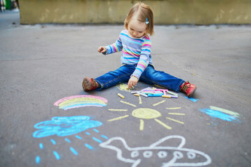 Adorable toddler girl drawing with colorful chalks on asphalt