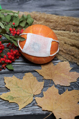 Orange pumpkin in a medical mask. Lies on black pine boards. Nearby is a bouquet of dried grass and mountain ash. Nearby are dried maple leaves.