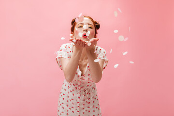 Joyful red-haired woman in sundress blows kiss on pink background with confetti