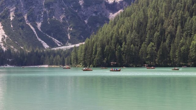 Lake Braies in summer, the emerald green water of the lake and the background of the forest with wild pine trees. People walk on the shore or row in boats on the surface of the lake.