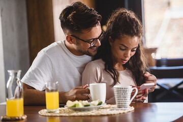 Beautiful interracial love couple using smart phone having good time at home in the morning after the breakfast