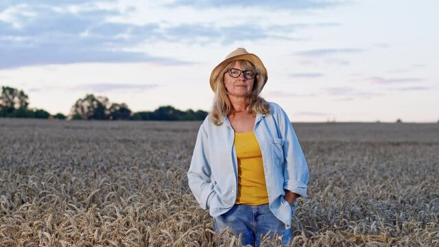 Image Of Beautiful Middle Aged Woman In Wheat Field. Beautiful Blonde In Hat And Glasses Holds Her Hands In The Pockets Of Jeans.