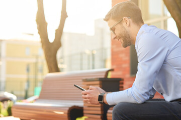 Chatting with friends. Young happy businessman wearing blue shirt and eyeglasses using smartphone and smiling while sitting on the bench on a sunny day