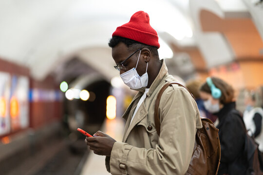 Black millennial man in trench coat, red hat wearing face mask as protection against  covid-19, flu virus, waiting for the train at subway station, using mobile phone. New normal, pandemic concept 