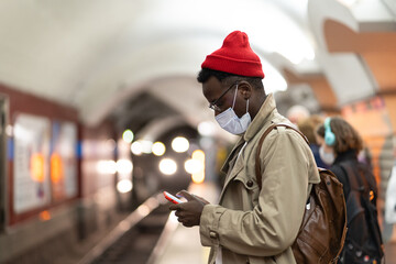 Black millennial man in trench coat, red hat wearing face mask as protection against  covid-19, flu...