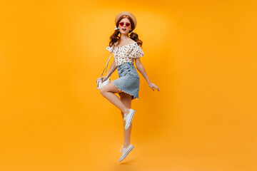 Fototapeta na wymiar Beautiful redhead girl in summer denim outfit and straw hat holding white bag and jumping on orange background