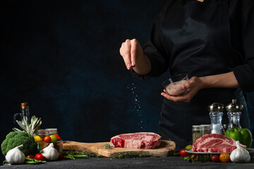 The chef in black uniform salts fresh pork steak on wooden chopping board on dark blue background. Cooking meat with spices at the professional kitchen. Frozen motion. Copy space on left.
