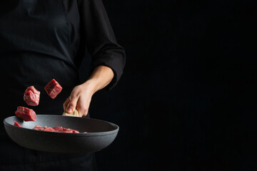 The hand of professional chef throws up meat cube in frying pan on black background. Concept of preparing food.