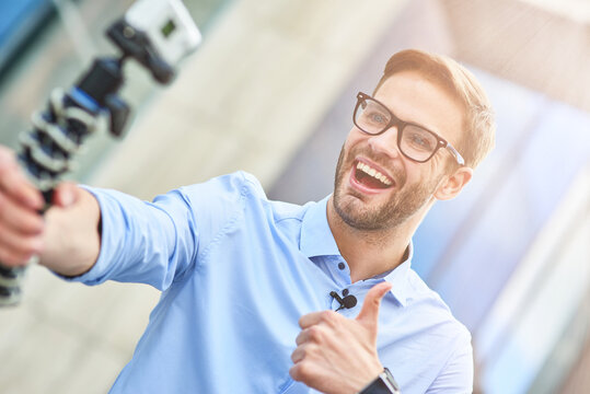 Young happy man, male blogger wearing blue shirt holding a gimbal with smartphone and recording video for his blog outdoors, smiling at camera and showing thumb up