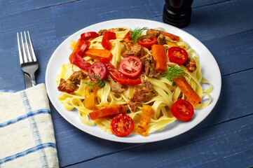 Pasta with vegetables. Fettuccine with grilled meat, carrots, bell peppers, dill tomatoes on a blue wooden background. Side view.