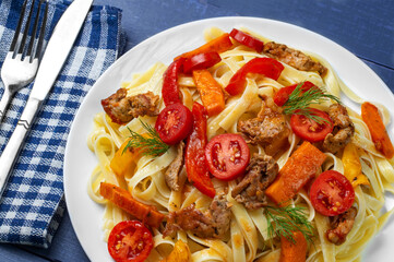 Fettuccine with grilled meat, carrots, bell peppers, dill tomatoes on a blue wooden background. Pasta with vegetables. View from above.
