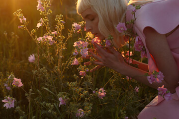 Girl with blond hair bent over flowers against sunset background . Portrait of girl with pink...