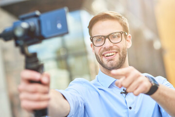 Young happy male blogger wearing blue shirt and eyeglasses holding a gimbal with smartphone, talking and smiling at camera while recording video for his blog outdoors