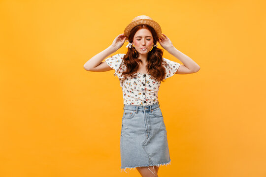 Snapshot of girl dressed in denim skirt and white blouse on orange background. Woman in boater inflates bubble of gum