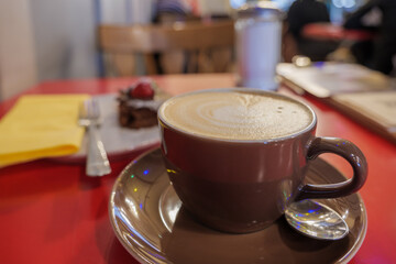 Selected focus and close up view of a brown ceramic cup of cappuccino on top with beautiful delicate latte art with heart pattern, and blur chocolate cake on side, on red table.
