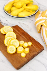 Sliced Yellow Crookneck Squash on a Wood cutting Board on Marble Countertop with a White Bowl of Uncut Squash in Background
